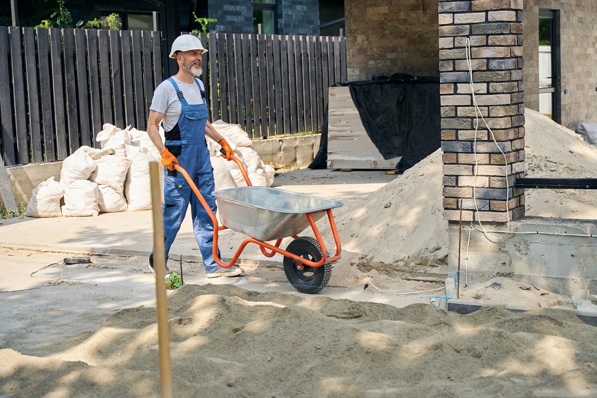 Worker in overalls and a hard hat pushing a wheelbarrow across a sand driveway base
