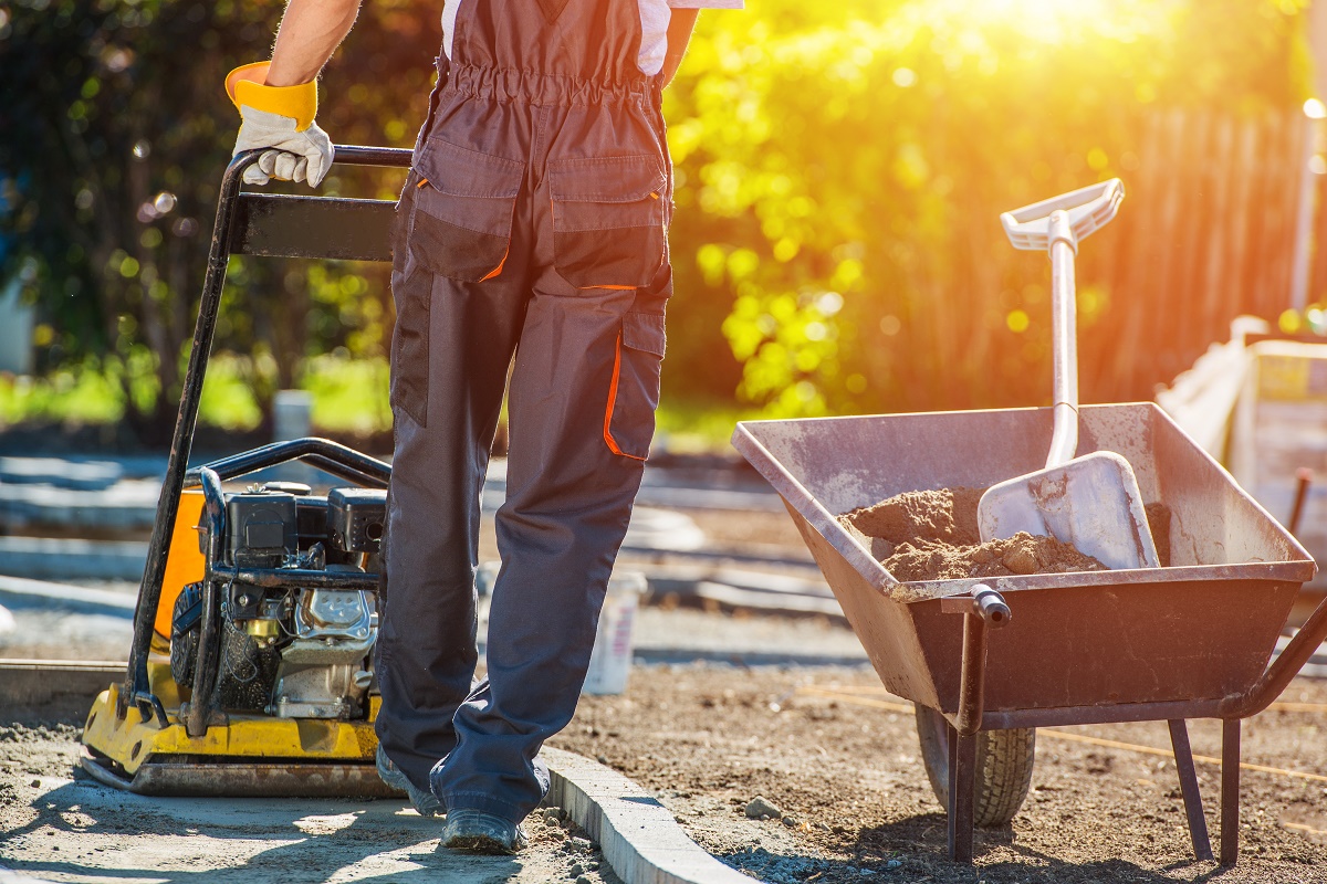 Worker running a plate compactor over a sand base beside a wheelbarrow