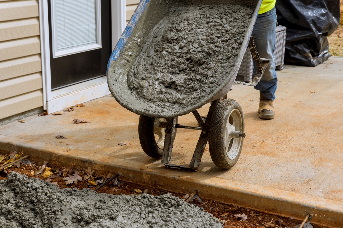 Worker tipping a wheelbarrow of wet concrete onto a driveway form beside a house
