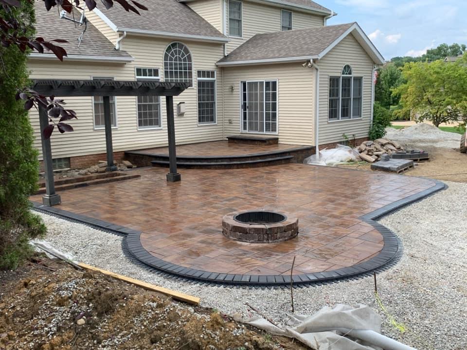 Large paver patio with a round stone fire pit and pergola under construction behind a house