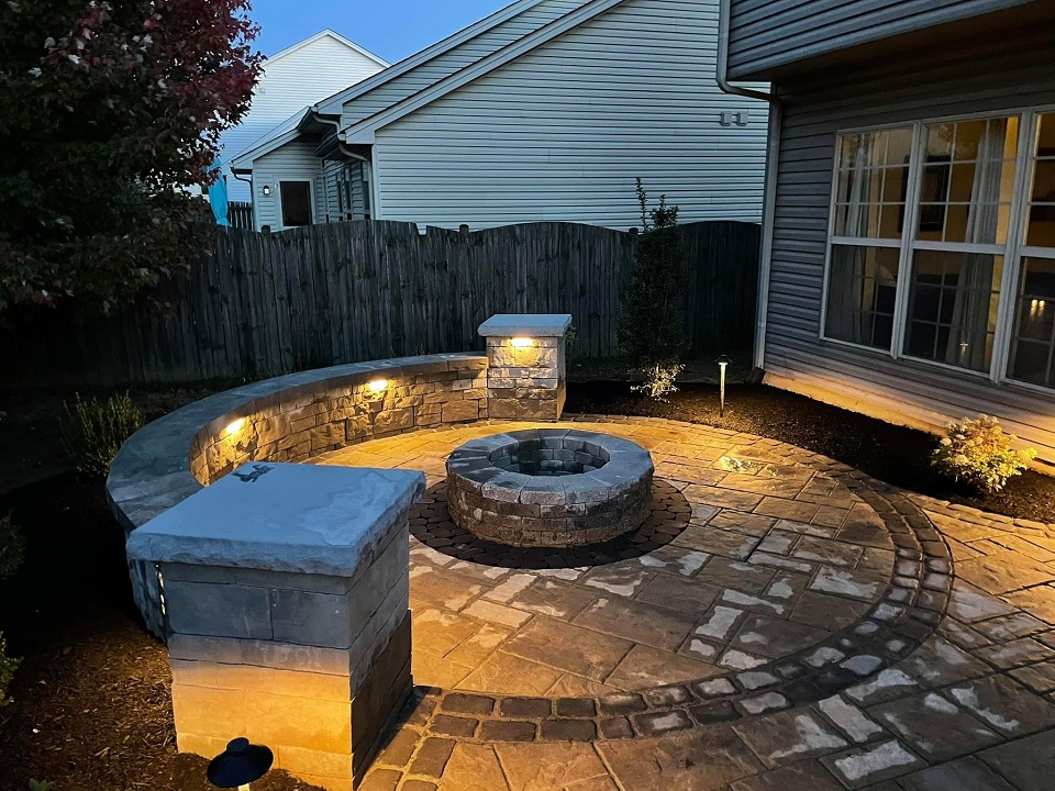 Circular paver patio at dusk with a stone fire pit, lit seat wall and capped pillars