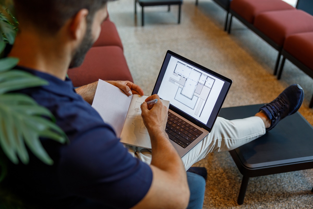 Man at a laptop viewing a CAD site plan while sketching in a notebook