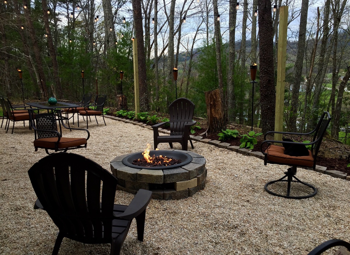 Gravel patio with a round stone fire pit, Adirondack chairs, string lights and tiki torches