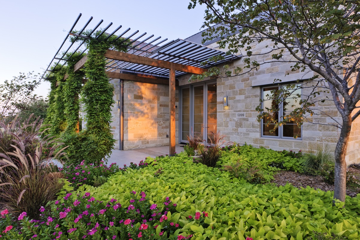 Stone house entry with a steel and cedar pergola over vines and annual flower beds