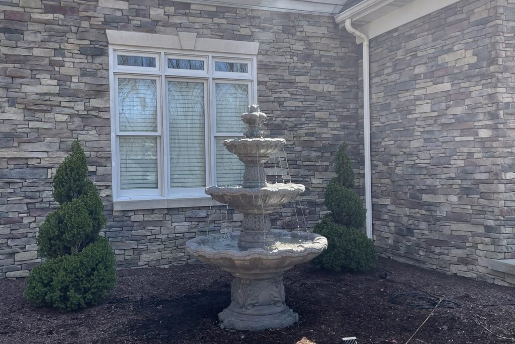 Tiered stone water fountain installed against a stone home with mulched beds and boxwoods by Daily Landscaping