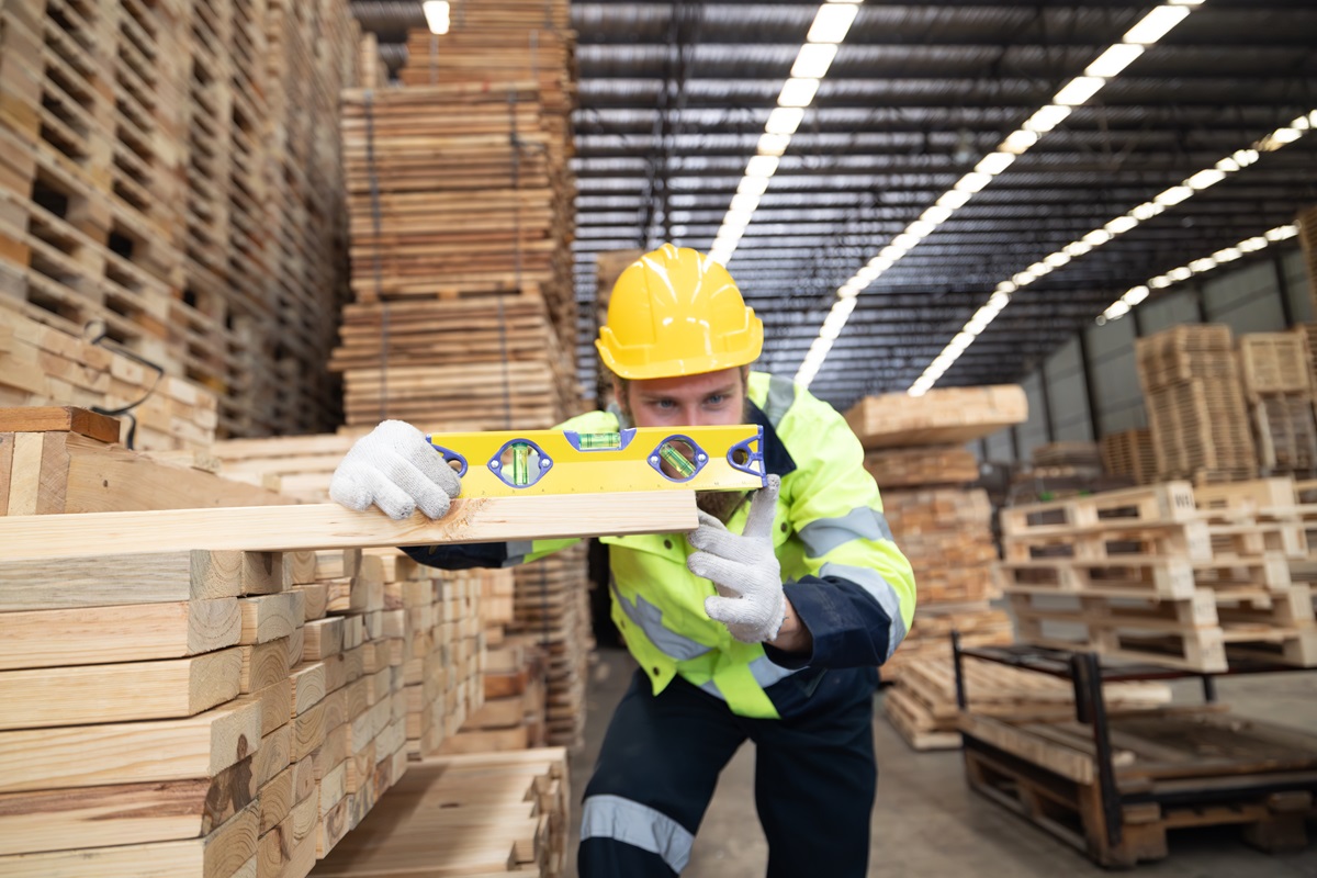 Worker in a yellow hard hat checking a lumber board with a level