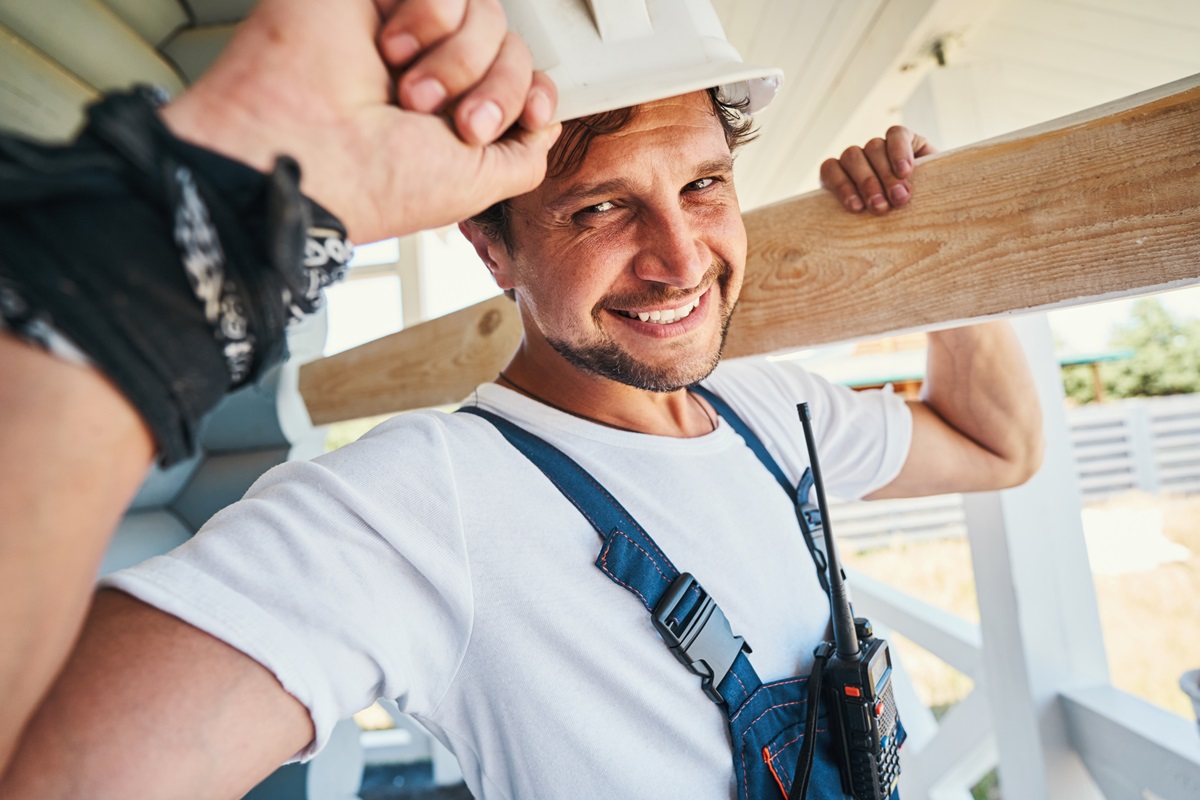 Smiling contractor in a hard hat carrying a lumber board on his shoulder
