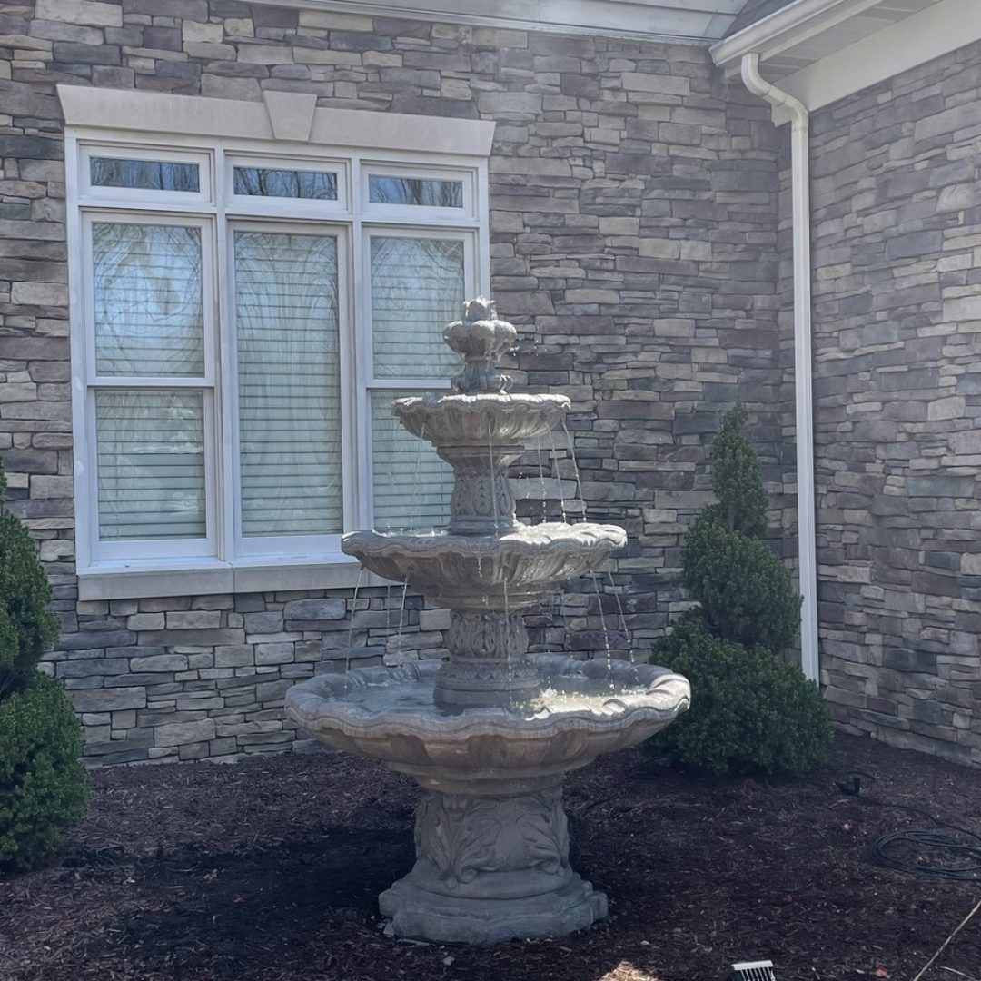Tiered stone water fountain installed against a stone home with mulched beds and boxwoods by Daily Landscaping